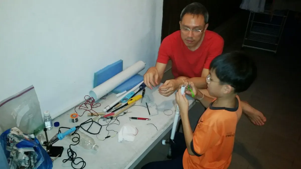 Kai as a schoolboy at a workbench, assembling a PVC air cannon out of pipe, tape and wire while an adult helps